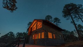 Long exposure of star trails swirling over a warmly lit wooden cabin surrounded by pine trees in a tranquil forest. Timelapse video - Powered by Shutterstock - Get 15% off with code: PIKWIZARD15