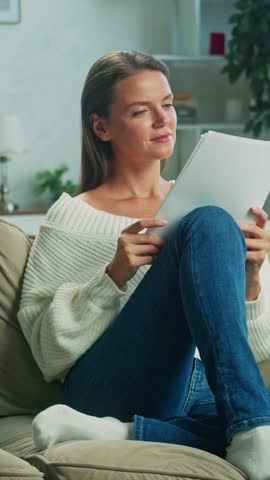 Woman sits comfortably on a sofa in her living room, leisurely reading through documents. The scene captures a peaceful atmosphere filled with natural light and tranquility