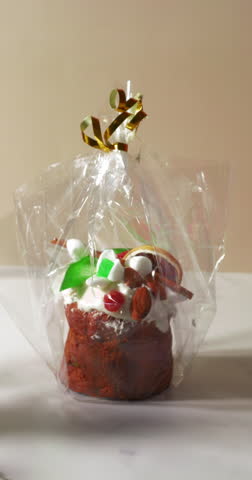 A festive mini fruit cake wrapped in clear cellophane with a gold ribbon, topped with frosting, dried fruit, almonds, and decorative sweets, ready as a holiday treat or gift.