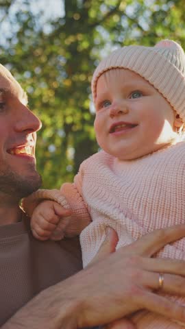 A father smiles warmly at his cheerful baby daughter, who is bundled up in a cozy outfit while being held in a park. Sunlight filters through the trees, creating a warm atmosphere.