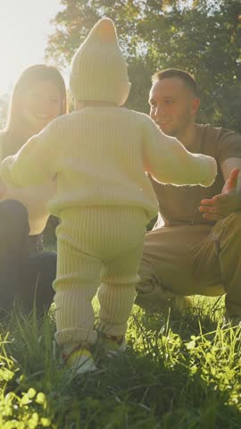 A joyful family spends a sunny afternoon in the park. A toddler in a cozy outfit playfully wanders toward smiling parents amidst the lush green grass.
