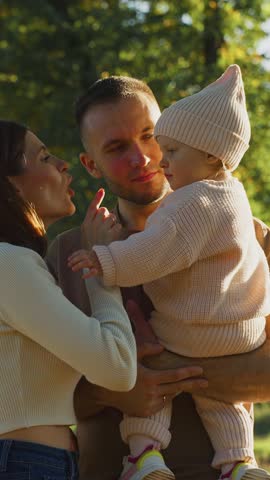 A couple stands in a park, with sunlight filtering through trees, as they engage playfully with their toddler. The mother playfully shushes the child while the father looks on lovingly.