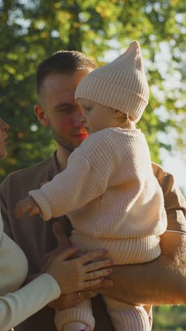 A couple stands in a park, with sunlight filtering through trees, as they engage playfully with their toddler. The mother playfully shushes the child while the father looks on lovingly.