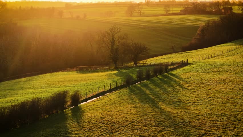 Golden light illuminating rolling farmland with wooden fences and scattered trees. Breathtaking farmland views and rolling hills in England at sunset. Warm sunlight casting long shadows over