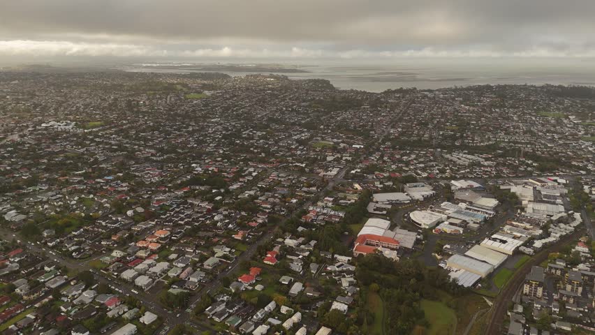 Wide aerial shot over Avondale suburb, Auckland, residential neighborhoods, commercial zone, harbour in distance under cloudy sky, suburbia. New Zealand. Drone pov