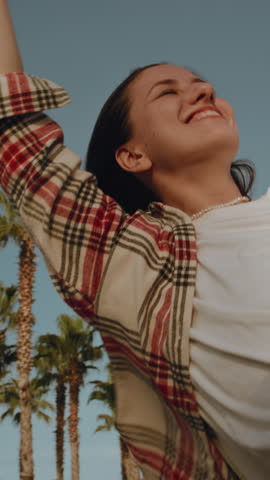 Vertical woman leans out the window of a rental car. Hair of girl is moving in the wind. Caucasian female is happy to see palm trees by sea or ocean on a beach, making waves with her hand. Road trip