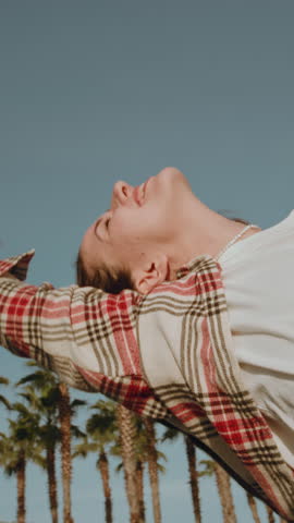 Vertical woman leans out the window of a rental car. Hair of girl is moving in the wind. Caucasian female is happy to see palm trees by sea or ocean on a beach, making waves with her hand. Road trip
