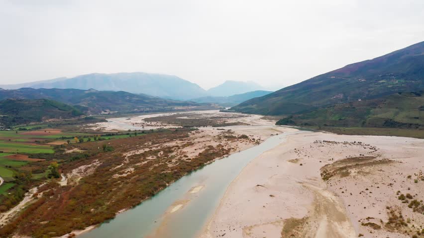 Aerial view of Albania’s Vjosa River, Europe’s last wild river. Turquoise waters carve through a pristine valley as the drone glides above this untouched natural wonder.

