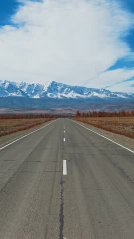 A long, open road stretches ahead toward breathtaking snow-capped mountains under a clear blue sky. The serene scenery invites exploration and adventure.