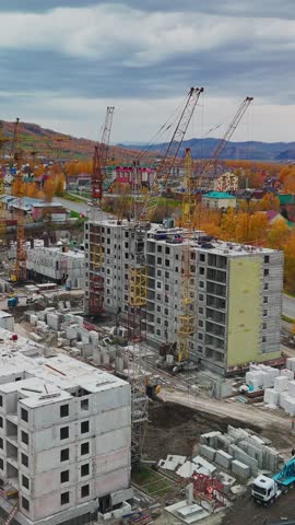 Workers and cranes are busy at a construction site in a neighborhood. New apartment buildings are taking shape amidst colorful autumn trees and a cloudy sky, showcasing urban development.