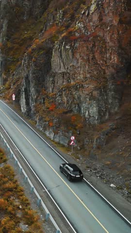 A black car navigates a winding road surrounded by rocky cliffs and vibrant autumn colors. The landscape showcases the beautiful natural scenery of the area, ideal for road trips.