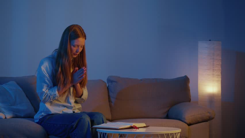 Young Woman Sitting Comfortably on Sofa in Evening Light, Hands Folded in Prayer. Peaceful and Calm Atmosphere of Quiet Prayer, Spiritual Reflection, Faith, and Personal Conversation with God.