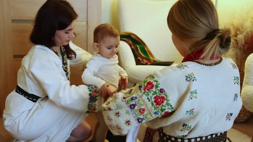 Two women dressed in traditional clothing assist a child in wearing a cultural outfit, showcasing heritage and family values in a warm, inviting environment.