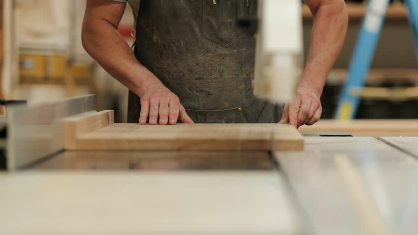 A skilled craftsman is operating a table saw, cutting a wooden plank with precision in a well-lit workshop. Dust particles float in the air, highlighting the craftsmanship involved.