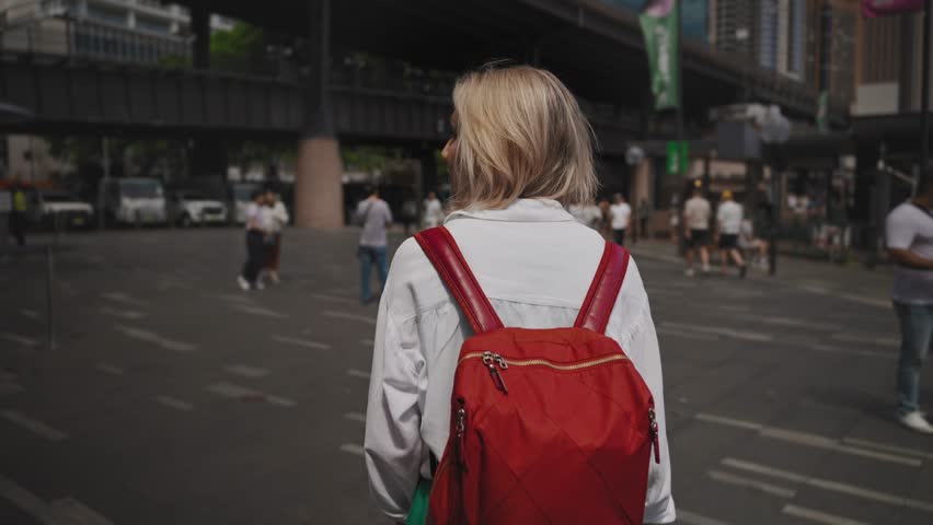 A young woman walks through a bustling city square on a summer day, captured from behind. This urban snapshot showcases everyday metropolitan life with dynamic crowds and architectural grandeur.