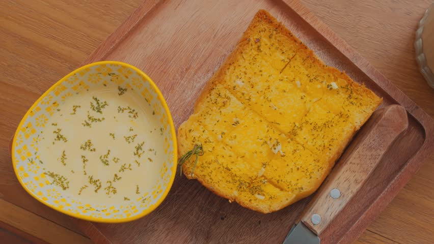 A wooden tray featuring a slice of golden, toasted bread topped with herbs and spices, alongside a small bowl of creamy dip or sauce with herbs sprinkled on top. A butter knife rests on the tray