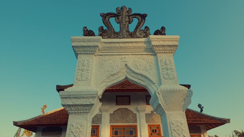 A beautifully carved white stone archway with intricate details and a crown-like structure at the top, set against a clear blue sky.