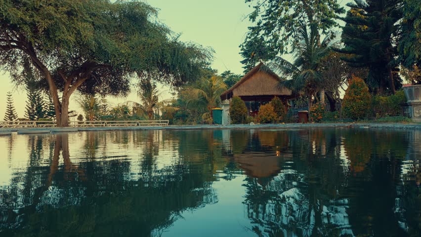 A serene landscape featuring a tranquil pond reflecting lush greenery and a rustic wooden house. Surrounding trees and plants enhance the peaceful atmosphere, creating a picturesque scene.