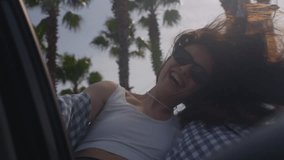 Excited woman in the window of a rental car, hair moving in wind. Caucasian female is happy to see palm trees by sea on beach. Hipster girl in shirt makes waves with her hand, goes surfing - Powered by Shutterstock - Get 15% off with code: PIKWIZARD15