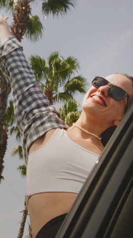 Vertical: Young woman performs dance movements leaning out of car window traveling along road, palm trees. Relaxed girl in sunglasses with pleasant smile revels in tranquility of moment, summer day