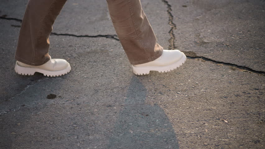 Young woman steps in white boots across cracked asphalt pavement under warm sunlight, winter pants swaying in rhythm, tapping echoes through empty urban street lined with melting snow
