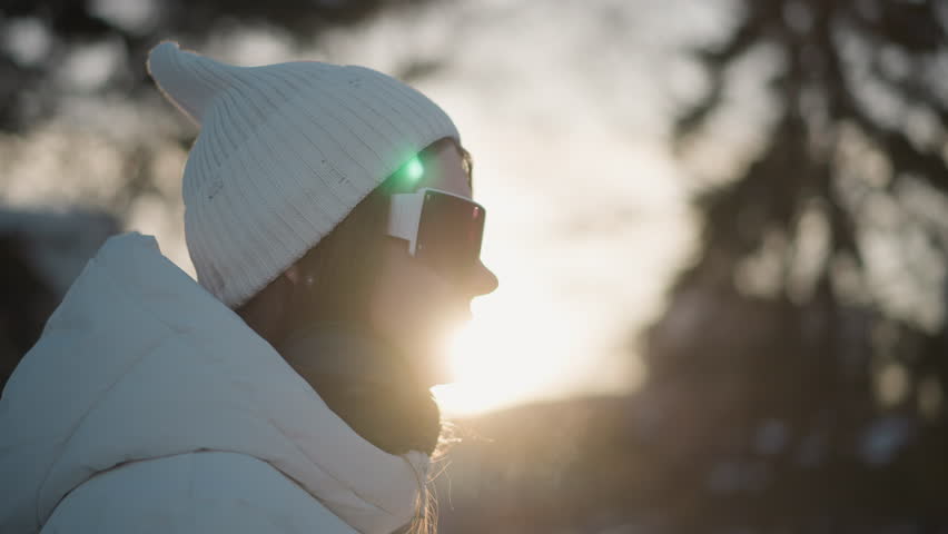 Side view of young girl holding phone to ear talking and smiling while shaking body under warm sunset light wearing white beanie and puffer coat against snowy forest background