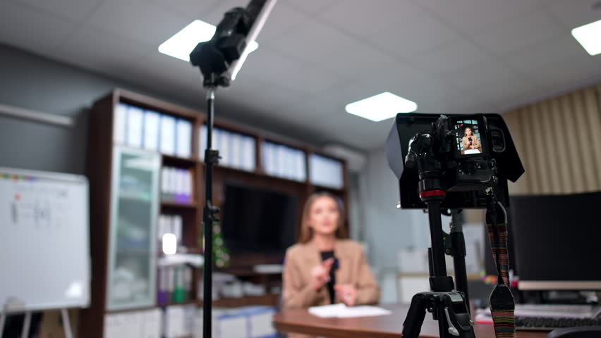 Day-light lamp on the stand and camera on tripod are set in the office. Woman in beige jacket recording blog content. Blurred backdrop.