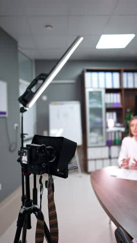 Approaching a modern camera on tripod filming a female speaker. Day light lamp stands near on the stand. Blogging concept. Blurred backdrop. Vertical video.