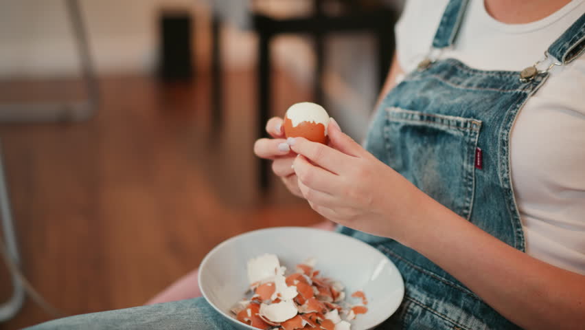 Portrait view of person wearing denim overalls carefully peeling egg shell over bowl filled with broken shells, with blurred background showing metal stand and home interior elements