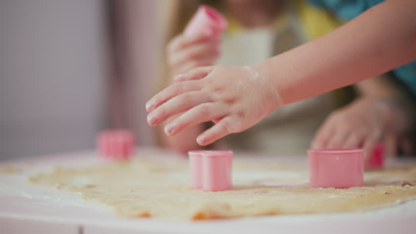 Close up of kid hand pressing pink cutter into flattened dough to create shapes, another kid blurred in background holding pink cutter, baking activity with flour spread over table