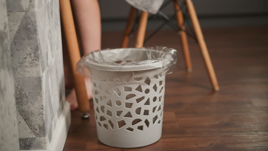 Ash colored patterned waste bin lined with plastic bag as person seated on wooden chair disposes potato peels into it, visible bare feet and wooden floor in cozy indoor setting