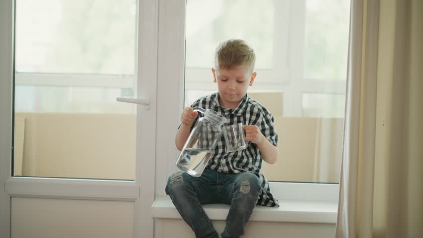 young boy wearing checkered shirt seated barefoot on window ledge pouring clear water from transparent jug into glass in bright indoor room with soft curtains and natural daylight