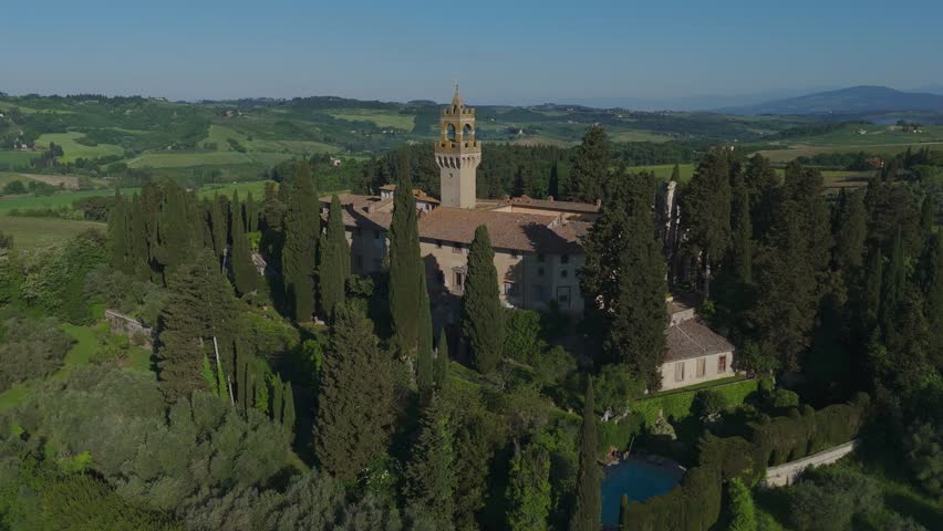 Drone shot approaching Castello di Montegufoni in Tuscany. Surrounded by cypress trees and vineyards, the castle emerges beautifully in the lush green Tuscan landscape under clear morning skies.