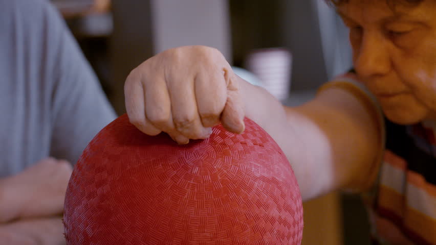 Middle-aged male caregiver gently assists the hemiplegic right hand ball therapy of a senior female stroke survivor, seated by glass top kitchen table at home. Back, and forth hand motion. 24fps.