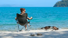 A woman feeds ducks by the sea. Redhead woman relaxing on camping chair, feeding ducks near ocean shoreline during bright summer day with peaceful seascape in background - Powered by Shutterstock - Get 15% off with code: PIKWIZARD15