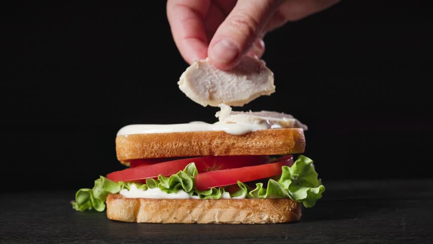 A cook put chicken meat on fresh toasted bread slice on black wooden board for making club sandwich, food closeup. Process of making healthy meal