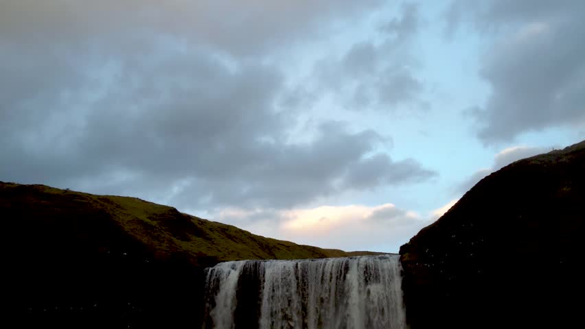 Aerial footage of yellow-toned mountains with a large waterfall in front and a lush green valley below. Wild and vibrant Icelandic landscape.