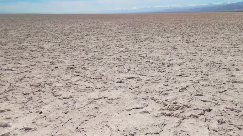 Drone footage flying forward over the Atacama Desert salt flats during daylight. The aerial view captures salt crusts and lithium deposits, revealing the arid landscape the horizon and the mountains