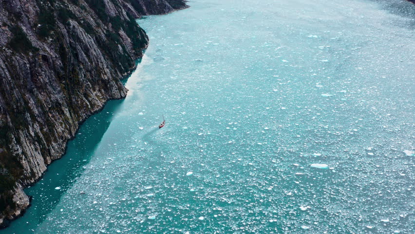 Aerial high angle of sailboat navigating turquoise Beagle Channel waters beside dark cliff face and foamy shoreline