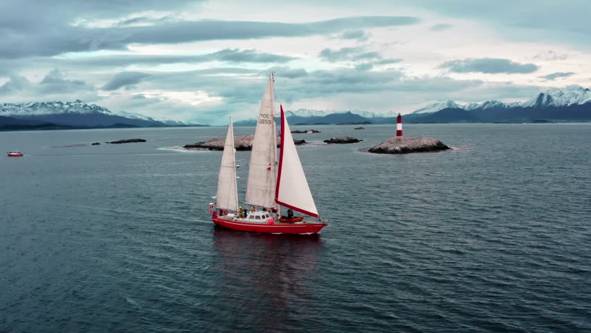 Red sailboat sails solo on wide Patagonian channel with gray sky and mountain background, aerial tracking right