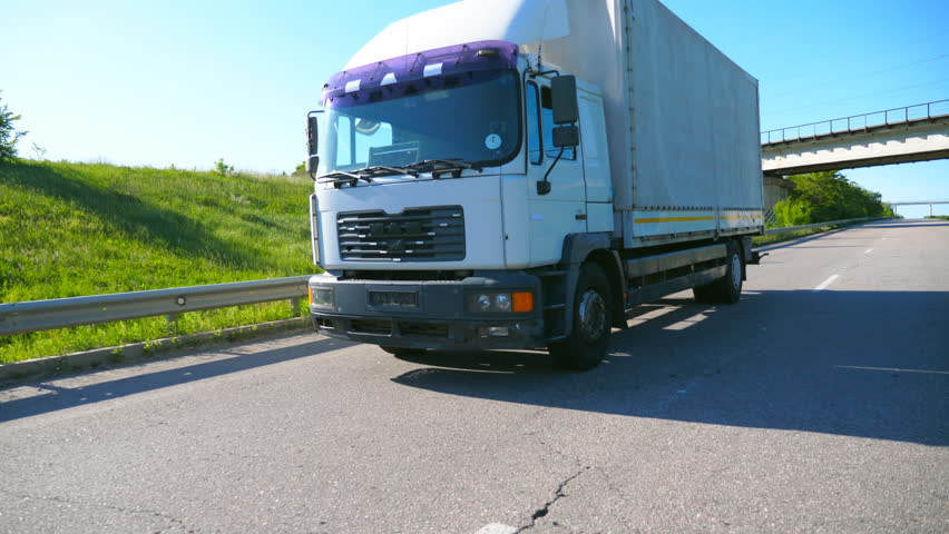 Big truck with cargo trailer driving on highway and transporting goods at sunny day. White lorry riding to destination through countryside road. Transport concept. Slow motion Side view Close up