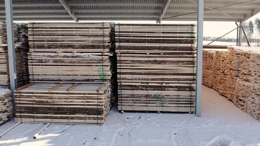 Neatly stacked lumber planks stored under a metal shelter, partially covered in snow, in a wintery outdoor lumber yard. No visible logos or people present