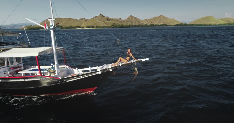Aerial drone footage of a young tourist relaxing and sunbathing on the bowsprit of a yacht sailing in the beautiful Komodo National Park, Indonesia