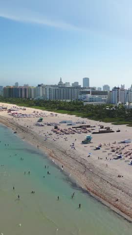 Vertical aerial 4K view above the coastline of Miami Beach, Florida, revealing a very busy shore packed with beachgoers, colorful umbrellas, and turquoise ocean waters under a tropical blue sky.