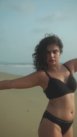 Woman in black bikini holds a poised dance stance with extended arms on a sandy beach by the sea