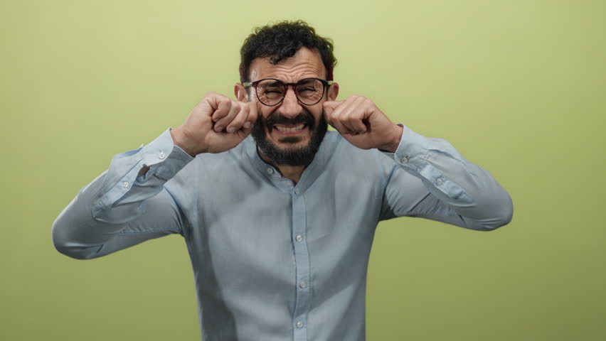 Hispanic man with beard and glasses mimics sadness against a yellow background, expressing emotions with hands near face, wearing blue shirt.