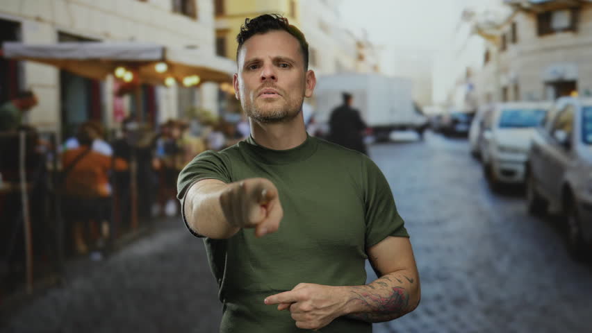 Man in green shirt with tattoos pointing on lively urban street with blurred people and vehicles in background depicting focus and interaction outdoors.