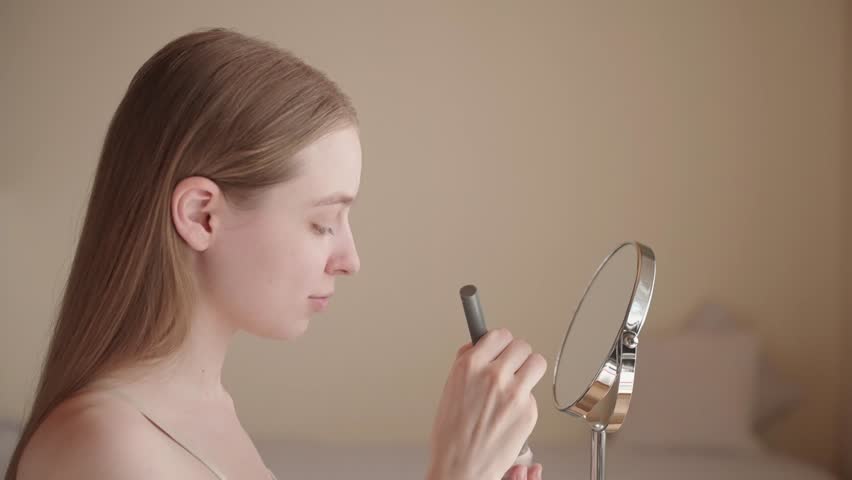 Cheerful young woman applying makeup with a brush as part of her morning beauty routine. A happy Gen Z female enjoying self-care and preparing for the day. Natural lighting, warm and calm atmosphere.