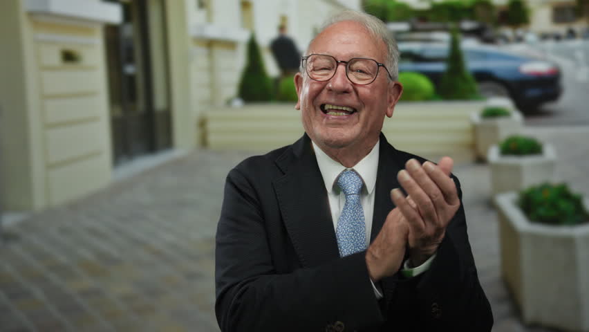 Senior man in a business suit clapping on an outdoor city street, exuding a joyful expression amidst urban surroundings and modern architecture.