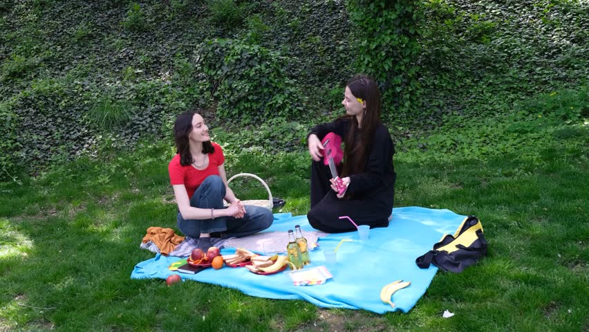 Young women enjoy picnic on light blue blanket in park playing ukulele and listening. Fruit drinks and wicker basket suggest picnic on green grass